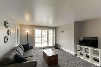 A modern and cozy living room featuring a dark gray sectional sofa, a rustic wooden chest coffee table, and a flat-screen TV on a white entertainment unit, with a sliding glass door revealing a snowy outdoor view.