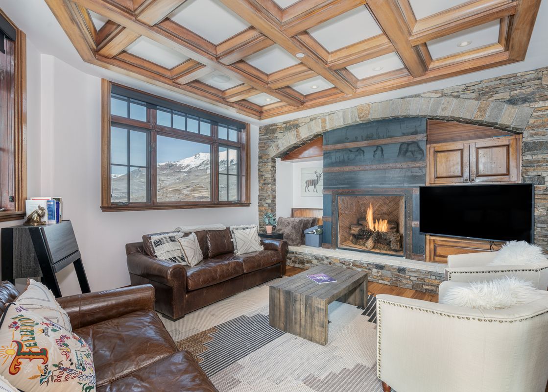 A cozy living room with a brown leather sofa, stone fireplace, and a large window offering mountain views at See Forever San Sophia 114 3BR 4BA.