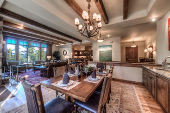 A luxurious dining and living area with rustic wooden furniture, exposed beams, and a large window overlooking a forest.