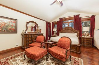 A luxurious bedroom at the Chateau at Timber Trail features a large wooden bed with white bedding, flanked by two nightstands with lamps, a dresser with a mirror, and two red patterned armchairs with an ottoman on an area rug, all under a vaulted ceiling