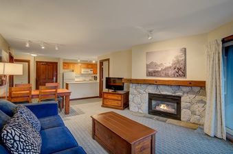 The living area of Blackcomb Springs One Bedroom Slopeside features a blue sofa, a wooden coffee table, a stone fireplace, and a dining table, with a kitchen visible in the background.