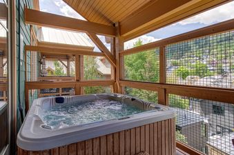 An outdoor hot tub with bubbling water on a wooden balcony, offering scenic views of lush greenery and distant houses under a clear sky.