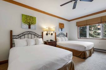 A hotel bedroom featuring two queen beds, each with white linens and pillows, separated by a wooden nightstand with lamps, under framed tree artwork.