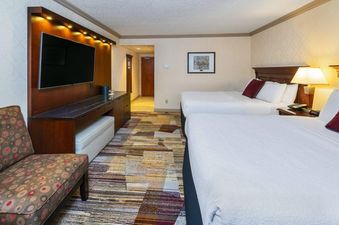 A hotel room with two queen beds, an entertainment unit with a flat-screen TV, and an accent chair.