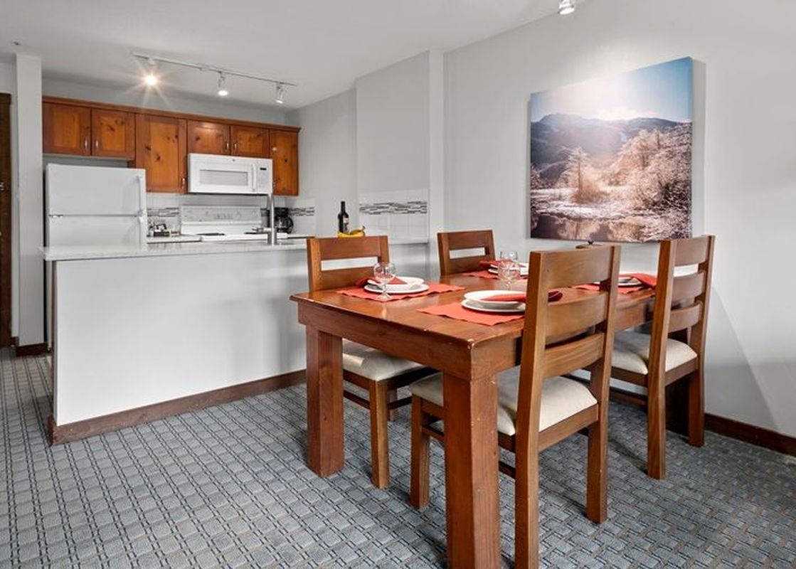 A dining table set for four with red placemats, adjacent to a compact kitchen with light wood cabinetry and white appliances.
