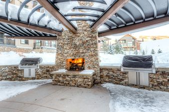 An inviting outdoor living area at Trailhead Lodge 2105 features a stone fireplace with a roaring fire and two barbecue grills, all surrounded by snow-covered grounds and buildings, perfect for winter entertaining.