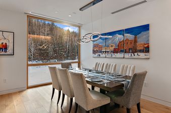 A modern dining room in Founders Place 3103 with a large window offering a winter mountain view, a dark wood dining table, upholstered chairs, and a colorful triptych art piece on the wall.