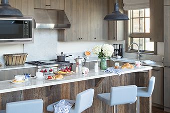 A rustic yet modern kitchen with wooden beams, light wood cabinetry, stainless steel appliances, a white granite island, and grey breakfast bar stools.