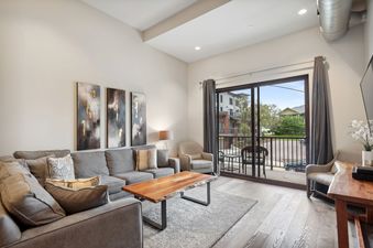 The image showcases the spacious living area of 2nd Street Lofts 209, complete with a large gray sectional sofa, a live-edge coffee table, abstract wall art, and a balcony with an exterior view.