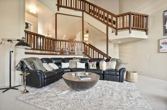 A spacious living room at Blue Bird on Norfolk featuring a large black leather sectional sofa, a round coffee table, and a faux fur rug, with elegant wooden staircases in the background.