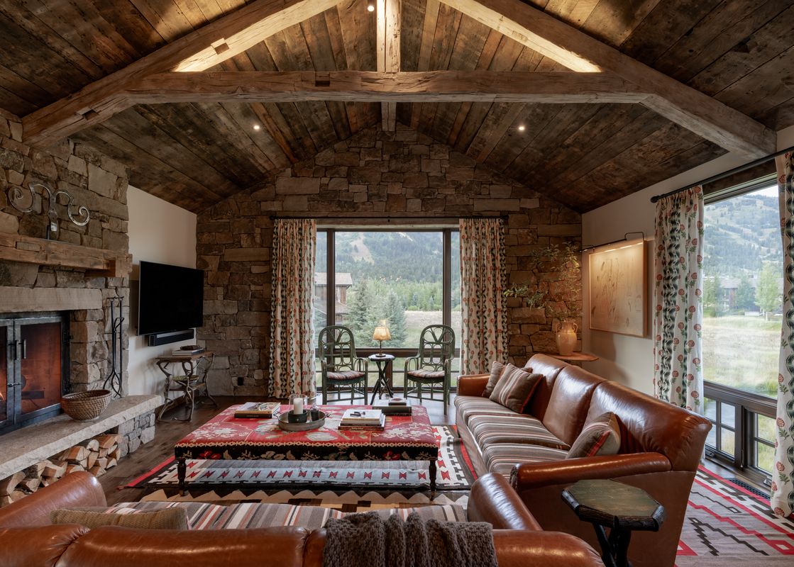 A cozy living room in Four Pines 09 with a stone fireplace, brown leather sofas, and a large window overlooking a scenic mountain landscape.
