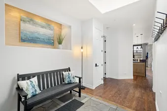 A welcoming entryway with a dark wooden bench and decorative pillows, an abstract blue and green painting in a recessed wall feature, and warm lighting leading to other rooms.