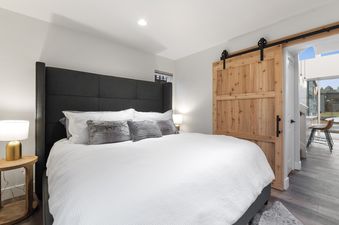 The comfortable bedroom at Quarry A28 features a king bed with a dark gray upholstered headboard, white bedding, and two side tables with lamps. A barn door leads to the rest of the unit.