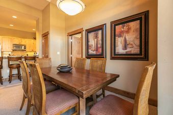 A dining table with six chairs is set up in a living room, there are two framed pictures above the table, the kitchen is visible in the background.