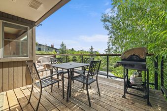 Private wooden deck at Snowridge 6 with a black dining table, chairs, a BBQ grill, and views of surrounding greenery.