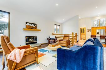 The bright and spacious living room at Snowy Peak Sanctuary features a blue velvet sofa, leather armchairs, a fireplace with mountain art, and large windows with outdoor views.