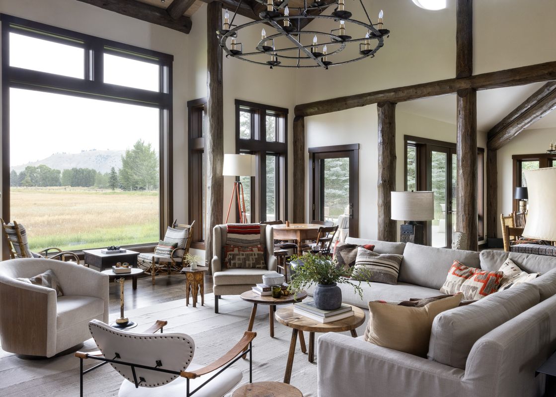 A rustic yet elegant living room with a large sectional sofa, accent chairs, and wooden coffee tables, all set against a backdrop of expansive windows showcasing a scenic outdoor landscape.