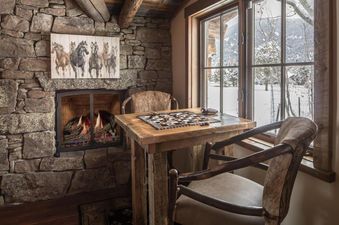 A cozy seating nook with a fireplace, a small wooden table, and two chairs by a window looking out onto a snowy landscape.