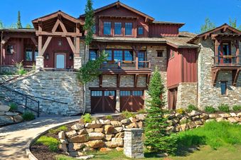 A large stone and wood house nestled among trees, with a winding driveway and retaining walls. The structure features multiple levels, balconies, and large windows, reflecting a harmonious blend of natural materials.