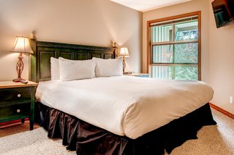 A cozy bedroom featuring a king-sized bed with white bedding, dark wooden furniture including a headboard and nightstands, and two lamps, with a large window offering a view of lush greenery.