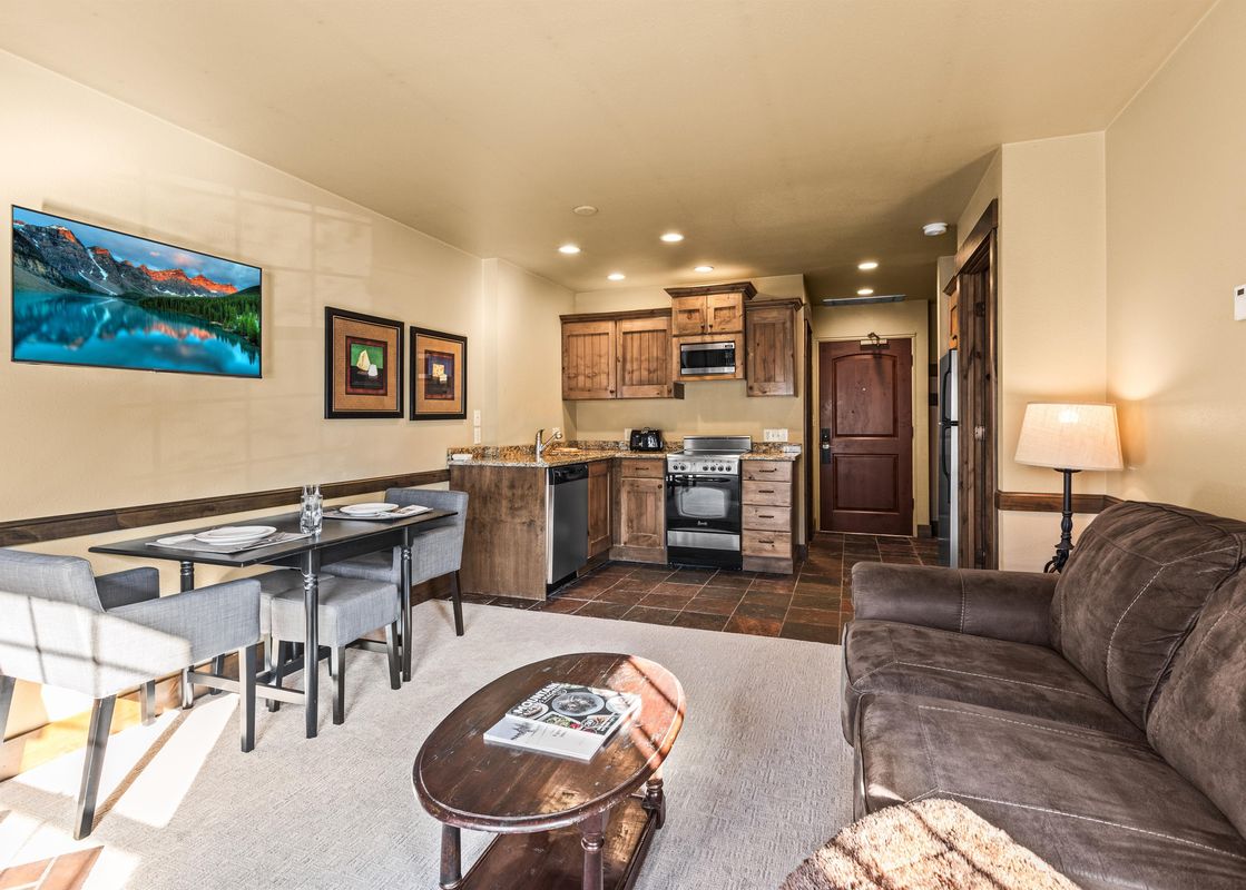 The living area and kitchen of Copperbottom Inn 102, featuring a brown sectional sofa, a coffee table, a dining table set for four, and a wall-mounted TV.