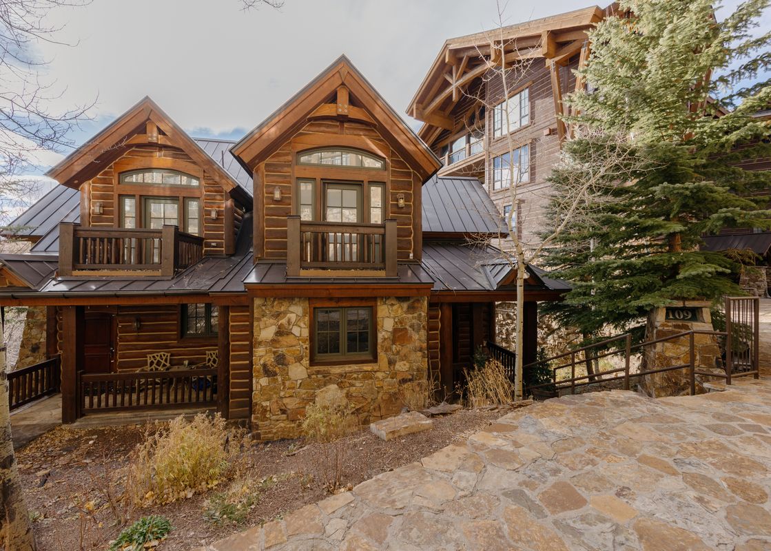 The exterior of Red Hawk Retreat at See Forever showcases a grand lodge-style home with log and stone construction, multiple balconies, and a dark metal roof, surrounded by natural landscaping and leading to a stone pathway.