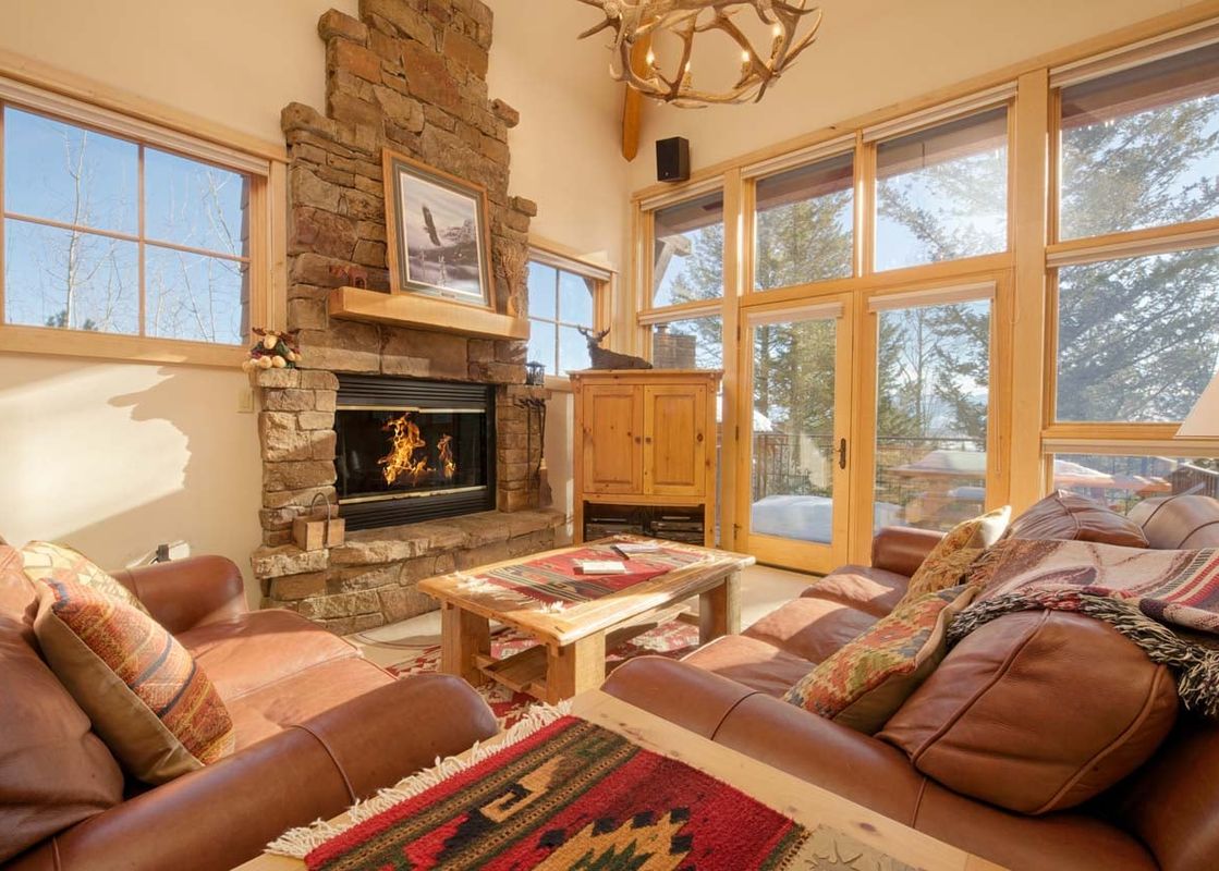 A cozy, sunlit living room at Moose Creek 31 featuring leather sofas, a large stone fireplace, and floor-to-ceiling windows looking out onto evergreen trees.
