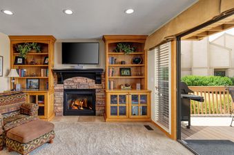 A cozy living room with a fireplace, two wooden bookcases, a patterned armchair, and a sliding glass door leading to an outdoor deck with a barbecue grill.