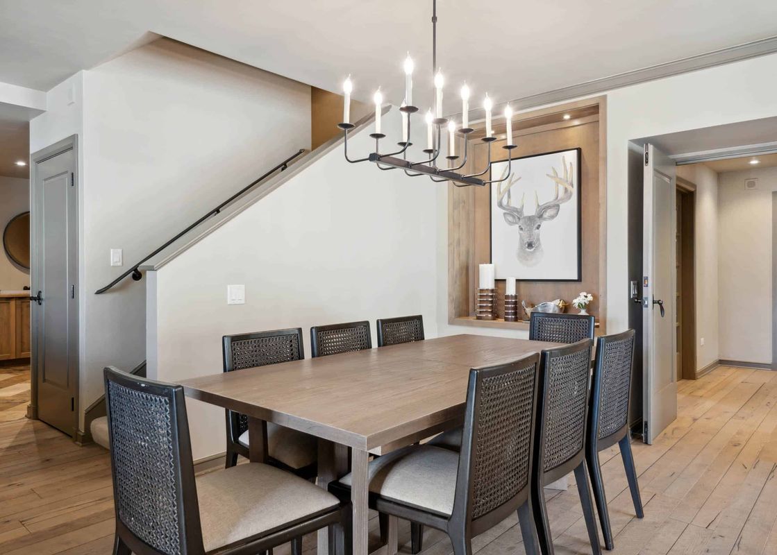 The dining area features a large wooden table and eight chairs with woven backs, lit by a stylish black chandelier, with a recessed wall niche displaying deer head artwork and decorative candles.