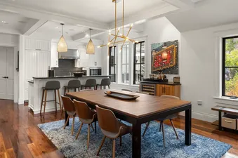 A modern dining area with a wooden table, stylish chairs, and a gold chandelier, seamlessly connected to a white kitchen with a dark countertop and bar stools, all set on a vibrant blue rug with large windows in the background.
