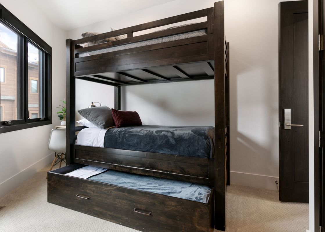 A modern bunk room with a dark wood bunk bed, trundle, and a large window offering an exterior view.