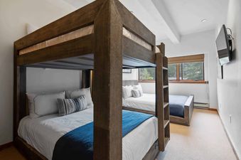 A bedroom with a wooden bunk bed, a full-size bed, and a window with a view of the trees outside