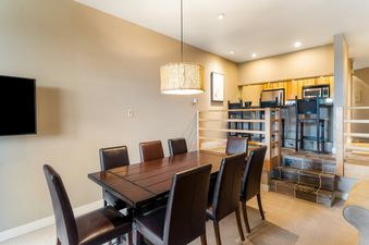 A dining room with a wooden table and leather chairs, a kitchen with a bar and stools, and a living room with a couch.
