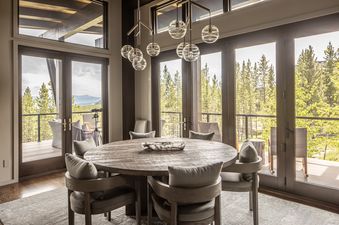 A rustic-chic dining room with a large round wooden table and plush chairs, overlooking a pine forest and distant mountains through expansive windows and glass doors.