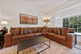 A cozy lounge area in The Charter B3305, featuring a large brown leather sectional sofa with decorative pillows, a modern black coffee table, and large bay windows overlooking green trees.