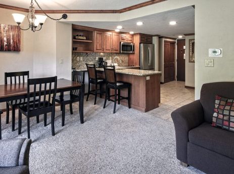 An open concept living area featuring a dining table with dark chairs, a kitchen with stainless steel appliances and a breakfast bar, and a cozy armchair.