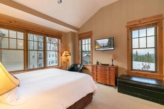 A bedroom with a large bed, a TV, a dresser, a black armchair, and windows with views of the mountains