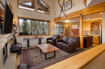 The living area of 12 Yukon Townhome features a large flat-screen TV above a fireplace, comfortable leather sofas and armchairs, and a coffee table, all set on a patterned rug. A large window offers stunning views of snow-covered trees, with the kitchen a