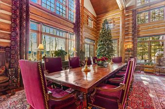 A wooden dining table with red velvet chairs, a Christmas tree, and a view of the forest outside of the window.