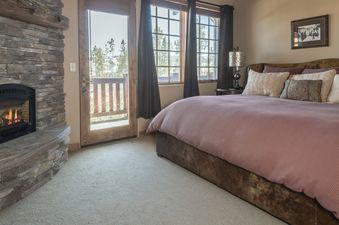 A cozy bedroom featuring a large bed with a pink duvet, a stone fireplace, and a glass door leading to a balcony with forest views.