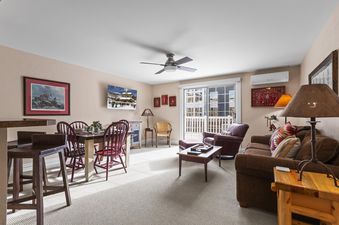 A bright living and dining area with light-colored walls, carpeted floors, and a mix of wooden and upholstered furniture.