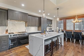 A modern kitchen with dark wood cabinets, white marble countertops, a black Viking range, and a kitchen island with gray bar stools. The dining area with an antler chandelier is visible in the background.
