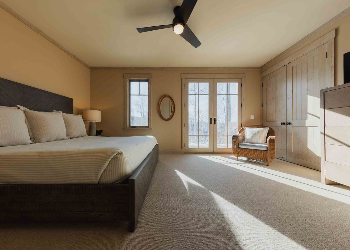 A cozy bedroom with a large bed, a wicker chair, and sunlight streaming through the windows.