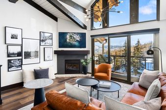 A spacious living room with a brown leather sectional sofa, a fireplace, and large windows offering a snowy mountain view.