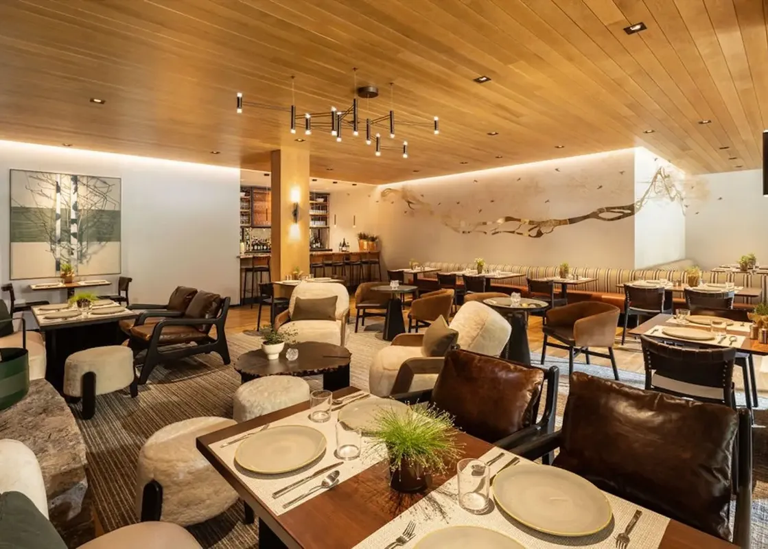 A modern and inviting dining area with wooden ceilings, stylish lighting, comfortable seating, and a bar area in the background.