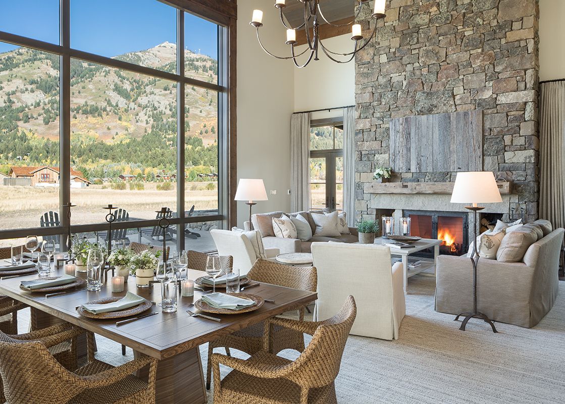 A lodge's great room showcasing a large stone fireplace, a formal dining area, and a cozy living space, all with expansive windows offering mountain views.