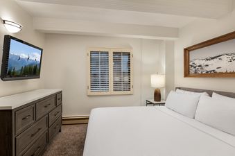 A hotel room with a white duvet bed, a dresser, and a window with white shutters.