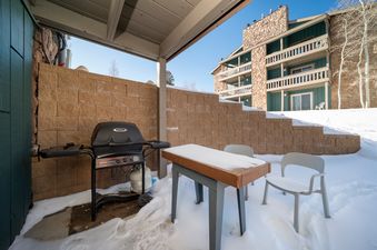 The outdoor patio of Silverado I C101, covered in snow, features a BBQ grill and a table with two chairs, set against a stone wall with a multi-story building in the background under a clear sky.