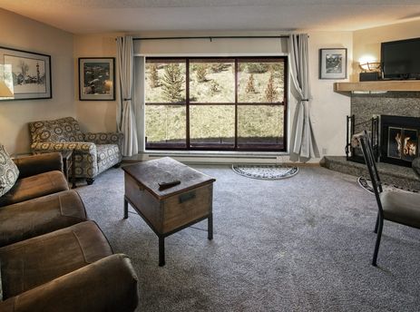 The living room of the Colorado Suite Beaver Run Resort is adorned with comfy sofas, a rustic coffee table, a grand fireplace, and a large window presenting a serene mountain view.