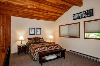 A cozy bedroom with a patterned bedspread, two nightstands with lamps, a wooden ceiling, and a 'MONTANA' sign above two windows.